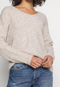 Beige ribbat stickad tröja som bärs med medelblå slitna jeans, händerna knäppta nära midjan, lockigt hår delvis synligt.