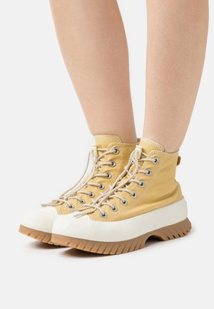 Zapatillas de caña alta en lienzo amarillo claro con puntera de goma blanca y suela gruesa y texturizada de color marrón. Presenta ojetes metálicos y cierre de cordones.