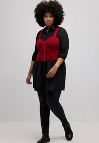 Red cropped top with a zipper and wide straps layered over a black button-up shirt, paired with dark jeans and ankle boots.