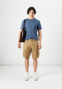 Casual outfit met een blauwe crew-neck t-shirt, kaki cargo shorts met zijzakken, en witte sneakers. Inclusief een brown schoudertas.