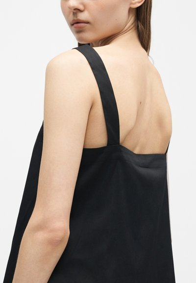 Femme à la peau claire portant une robe noire sans manches, vue de dos et au niveau des épaules, légèrement tournée vers la gauche sur un fond uni.