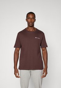 Brun bomulds t-shirt med rund halsudskæring, korte ærmer og et lille hvidt logo på brystet. Sammen med lysegrå sweatpants.