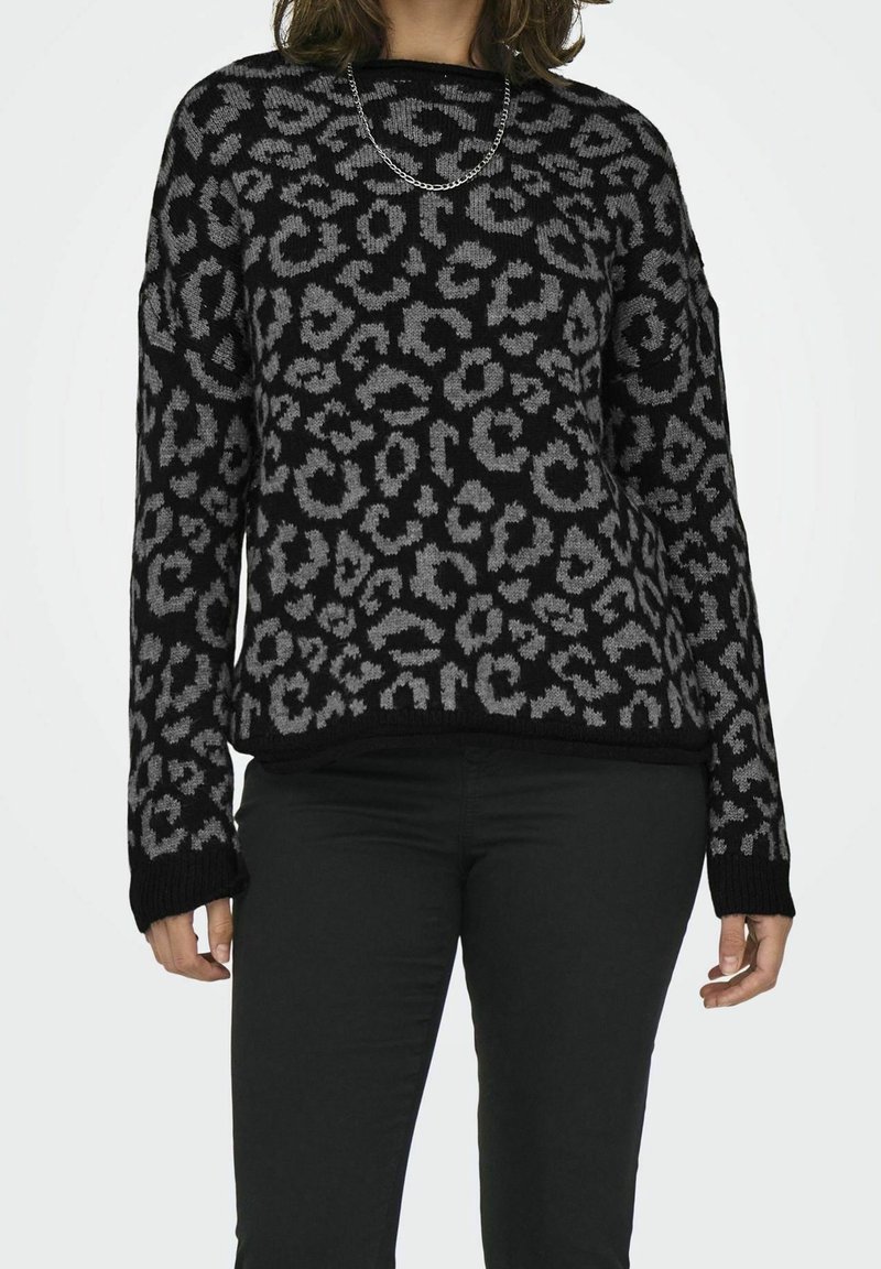 Personne portant un pull noir et gris à imprimé léopard, un pantalon noir et un court collier chaîne argenté sur un fond blanc.