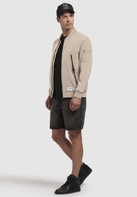 Beige bomberjakke, lynlåslukning foran, ribbede manchetter, sidelommer, båret over en sort skjorte, parret med mørke shorts og sorte høje sko.