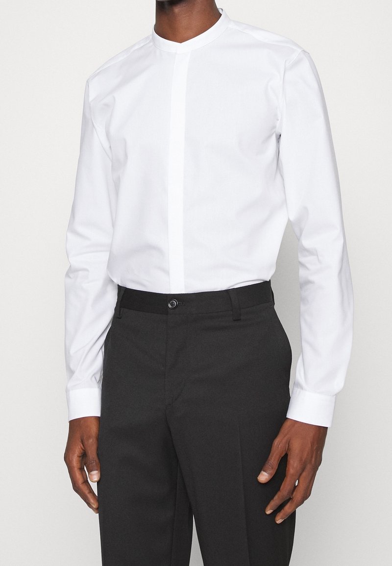 Homme portant une chemise blanche cintrée à col mandarin avec des boutons cachés et un pantalon de costume noir, debout devant un fond uni.
