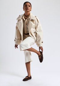 Beige trench coat dengan kerah besar, dipakai di atas blus bergaris dan celana putih model cropped. Sepatu dengan pola macan tutul coklat dengan tali.