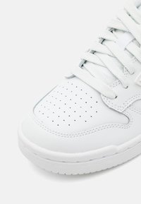 Bout de basket blanc en cuir perforé, lacets blancs, détails de couture et semelle en caoutchouc texturée sur fond blanc.