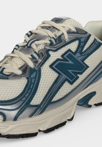 Крупен план на бяла и синя спортна обувка New Balance с мрежеста тъкан, връзки и текстурирана гумена подметка.