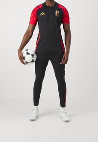 Schwarzes Sporttrikot mit roten Akzenten, Adidas-Logo, belgisches Flaggen-Patch, kombiniert mit schwarzen enganliegenden Hosen und einem Fußball, getragen mit Fußballschuhen.