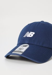 Navy blue baseball cap with embroidered white logo, curved visor, adjustable strap, and woven texture. Features label with care instructions.