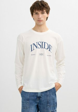 Joven con camisa blanca de manga larga con el texto "INSIDE" y jeans azules, de pie contra un fondo blanco liso.