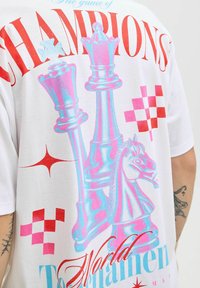 Baltā T-krekliņa priekšpusē ir liela šaha figūru grafika rozā un zilā krāsā, ar sarkanu tekstu "ČEMPIONU spēle" un punktveida ornamentiem.