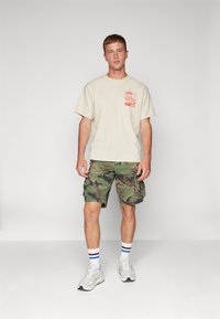 Beige graphic t-shirt with red design, green camo cargo shorts, white sneakers with gray accents, and striped socks in blue and red.