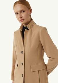 Beige Wolle-Mix-Mantel mit hohem Kragen, vorderem Knopfverschluss und seitlichen Taschen. Glatte Textur und taillierte Passform mit Nahtdetails.