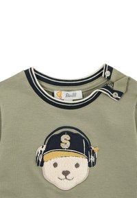 T-shirt verde oliva con colletto tondo, dotata di un design ricamato di un volto d'orso con cuffie e cappello. Bottoni a pressione sulla spalla.