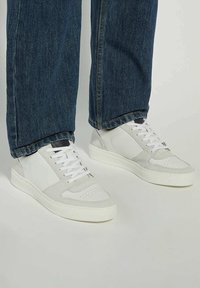 White athletic sneakers with suede and leather accents, flat laces, round toe design, and a textured rubber sole, paired with dark denim jeans.