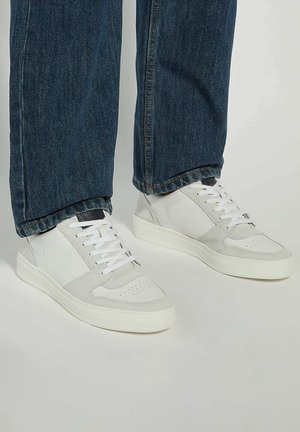 White athletic sneakers with suede and leather accents, flat laces, round toe design, and a textured rubber sole, paired with dark denim jeans.