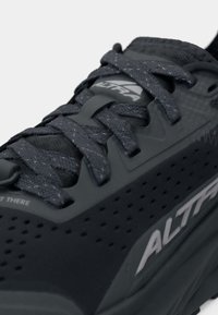 Shoe atlética cinza com superfície texturizada, cordões espessos e um design perfurado. Apresenta um logotipo e detalhes refletivos na lateral.