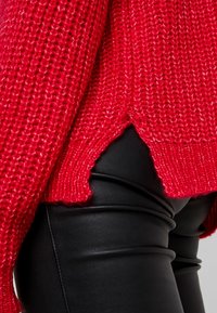 Röd stickad tröja med ett texturerat mönster och sidoklyftor, matchad med tajta svarta byxor i konstläder med horisontella sömmar.