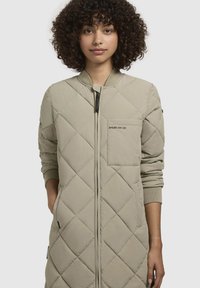 Giacca beige trapuntata con zip frontale, polsini a costine e tasca sul petto. Presenta un motivo a diamanti e una vestibilità rilassata.