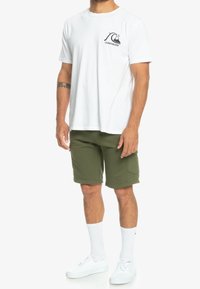 Bílá bavlněná trička s černým vlnovým logem na hrudníku, spárované se světle zelenými šortkami, bílými teniskami a bílými ponožkami.