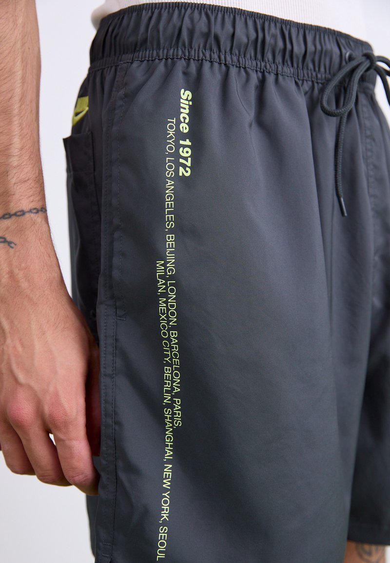 Costumi da bagno grigi con fascia elastica in vita e cordoncino. Presentano un testo verde che elenca le città e la dicitura "Dal 1972" sul lato.