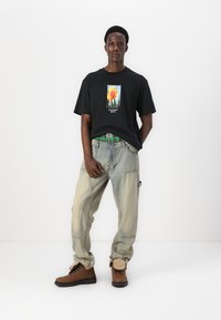 Tricou negru cu un grafic floral colorat, blugi deschiși și largi, și cizme maro cu accente bej. O ținută cu croială lejeră.