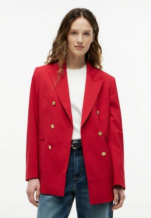 Frau mit welligem braunem Haar trägt einen rot-doppelreihigen Blazer mit goldenen Knöpfen, weißes Oberteil, dunklen Gürtel und blaue Jeans vor weißem Hintergrund.