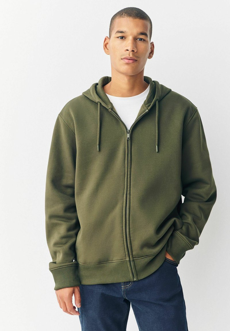 Junger Mann trägt einen olivgrünen Reißverschluss-Hoodie über einem weißen Hemd und dunkelblauen Jeans. Er steht mit einer Hand in der Tasche vor einem schlichten Hintergrund.