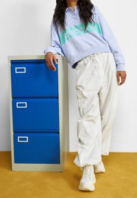 Ljusblå sweatshirt med grön accent och logotyp, vita cargobyxor och krämfärgade sneakers, bredvid en ljusgrå arkivskåp med blå lådor.