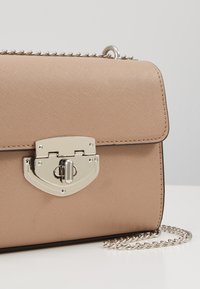 Beżowa torba z ekoskóry o fakturowanym wykończeniu, z elementami w srebrnym odcieniu, zamknięciem na klapę i łańcuszkowym paskiem. Wyróżnia się eleganckim wzorem zamka.