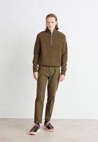 Pull en maille marron avec un col zippé mi-hauteur, associé à un pantalon vert olive et des chaussures noires avec des lacets roses et une semelle légère.