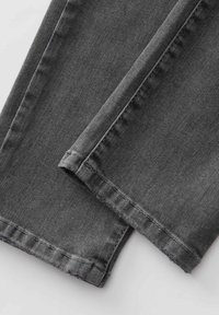 Primo piano di gambe di pantaloni di jeans neri con cuciture visibili e orli leggermente sfilacciati su uno sfondo chiaro.