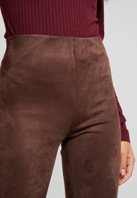 Leggings en daim marron avec une taille haute, présentant une texture lisse et une coupe ajustée, partiellement visibles à côté d'un haut bordeaux côtelé.