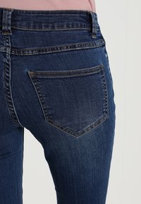 Tmavě modré džíny s přiléhavým střihem, které mají malou šikmou zadní kapsu a viditelné švy podél pasu.