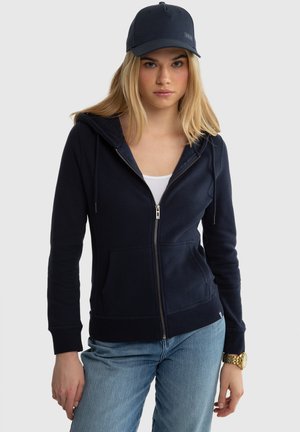 Mladá žena oblečená v námořnicky modré mikině na zip, světle modrých džínách, bílém tričku, námořnické čepici a zlatých náramkových hodinkách na jednobarevném pozadí.