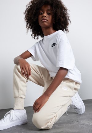 Camiseta blanca de Nike con logo negro, pantalones de chándal beige con logo blanco y zapatillas blancas. El conjunto presenta un ajuste holgado y material suave.