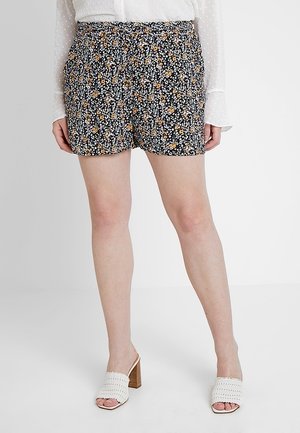 Kvinde iført sorte blomstrede shorts, hvid tekstureret bluse og hvide hælede flettede sandaler, stående mod en ensartet baggrund.