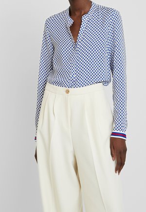 Persoon draagt een blauw-witte polkadot blouse met lange mouwen, ingestopt in een hooggewaisterde crème wijde broek met knoopsluiting.