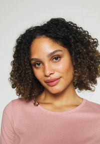 Roze lange mouwen top met een ronde hals, zachte textuur en een getailleerd ontwerp. Model heeft krullend haar, neutrale achtergrond.