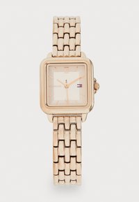 Montre à quartz avec un bracelet en métal rose doré et un boîtier rectangulaire. Présente un cadran clair avec des chiffres romains et le logo de la marque en bas.