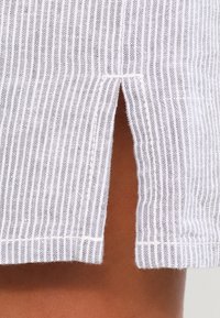 Gros plan d’un tissu blanc avec de fines rayures verticales grises et une petite fente centrale révélant une peau brune en dessous.