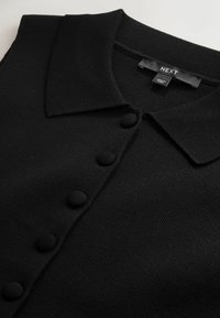 Juodas megztas drabužis su apykakle ir šešiais apvaliais juodais mygtukais priekyje. Matoma etikėtė su prekės ženklu „NEXT“. Tekstūruota medžiaga.