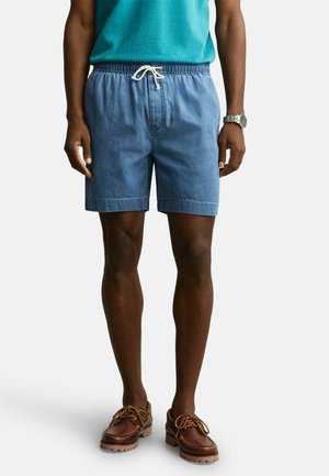 Man wearing blue drawstring shorts, brown leather casual shoes, turquoise shirt, and silver wristwatch, standing against a white background.