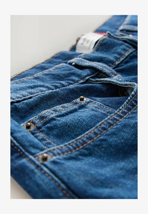 Jean en denim de couleur bleu foncé avec des coutures orange contrastantes, comprenant des poches et des rivets en métal aux coutures des poches. Tissu texturé visible.
