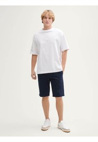 Weißes Baumwoll-T-Shirt mit lockerer Passform, kombiniert mit dunkelblauen Shorts. Die Accessoires bestehen aus weißen Sneakers mit hellbraunen Sohlen und minimalistischem Design.