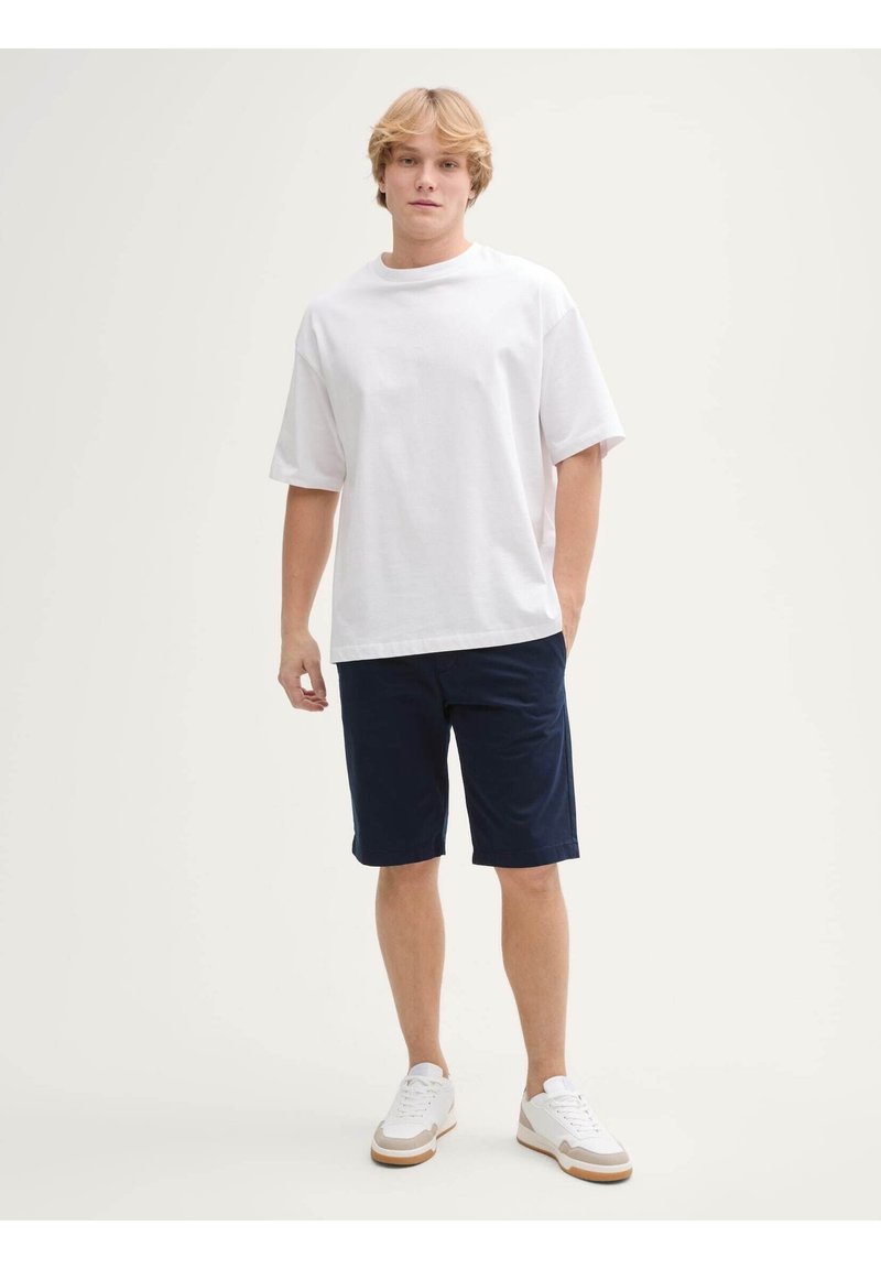 Weißes Baumwoll-T-Shirt mit lockerer Passform, kombiniert mit dunkelblauen Shorts. Die Accessoires bestehen aus weißen Sneakers mit hellbraunen Sohlen und minimalistischem Design.