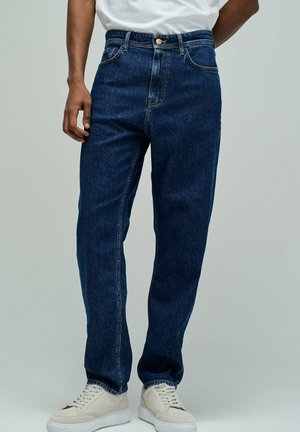 Hombre con vaqueros rectos azul oscuro y zapatillas blancas, de pie con una mano al lado de la cadera.