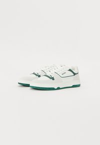 Hvite sneakers med grønne detaljer, med en glatt læroverdel, rund tå, snøring foran og teksturert gummisåle. "BORG"-logo på siden.