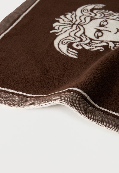 Καφέ πετσέτα με λευκό κεντητό σχέδιο κεφαλής Μέδουσας και υφασμένο περίγραμμα, διπλωμένη πάνω σε λευκή επιφάνεια.